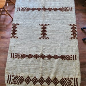 May include: Cream-colored, rectangular table runner with a rounded end. The runner features a repeating geometric pattern in shades of brown. The design includes diamond and rectangular shapes, creating a textured appearance. This textile is suitable for home decor.