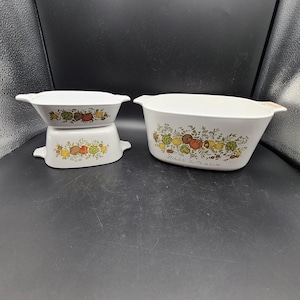 May include: A set of three white ceramic casserole dishes with a green, yellow, and red floral design featuring vegetables and the text "Le Chic de la Marjolaine".
