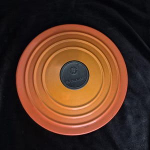 May include: A round, orange and yellow gradient enameled cast iron pot lid with concentric circular ridges. The lid features a centered, circular black knob with the embossed text LE CREUSET and a small logo in the middle of the knob surface.