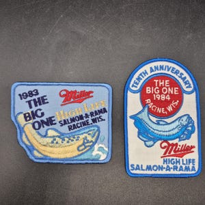May include: Two embroidered fabric patches. The left patch is light blue with a yellow fish and text reading 1983 THE BIG ONE MILLER HIGH LIFE SALMON-A-RAMA RACINE, WIS. The right patch is white with a blue fish and TENTH ANNIVERSARY THE BIG ONE 1984.