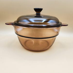 May include: An amber-coloured glass casserole dish with a matching lid. The dish has two side handles and a dark knob on the lid. The glass is transparent. A classic kitchenware item, perfect for cooking and serving.