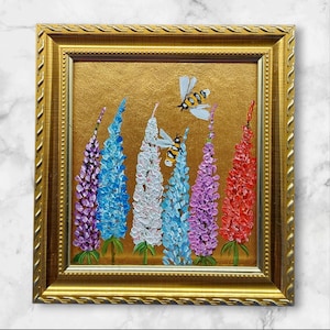 Op de afbeelding: Een ingelijst schilderij met kleurrijke lupinebloemen in tinten paars, blauw, wit en rood, tegen een gouden achtergrond. Twee bijen zijn afgebeeld die in de buurt van de bloemen vliegen. Het schilderij is ingesloten in een gouden, sierlijke lijst.
