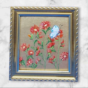 May include: A framed painting depicting red flowers and a light blue butterfly with white spots. The artwork is set against a gold background and framed in a gold and blue ornate frame. The painting is a floral theme.