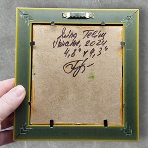 以下が含まれることがあります： 緑と黄色のフレームが付いた額装された絵画の裏側。絵画には署名があり、「Irina Telus Ukraine, 2024, 12.2cm x 10.9cm」というテキストが含まれています。フレームには鋸歯状のハンガーが付いています。