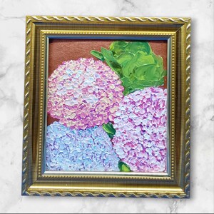 Op de afbeelding: Een ingelijst schilderij van roze en blauwe hortensia's met groene bladeren. Het kunstwerk staat tegen een roodbruine achtergrond en is goudkleurig omlijst. Het schilderij heeft een textuur, impasto-stijl. De lijst is sierlijk.
