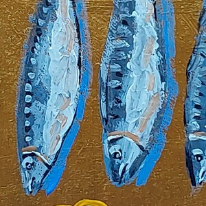 以下が含まれることがあります： 金色の背景に、青い魚が3匹描かれた芸術的な絵画。白とベージュのディテールが施されています。下部には黄色のレモンスライスがあります。この作品は、テクスチャのある塗装された外観をしています。