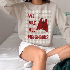 以下が含まれることがあります： 赤い文字で「WE ARE ALL NEIGHBORS」と書かれたベージュのスウェットシャツ。デザインには、青いネクタイが付いた赤いカーディガンが含まれています。スウェットシャツは衣料品コレクションの一部です。