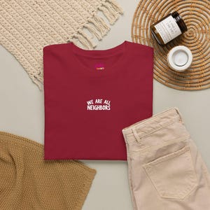 May include: A dark red crew neck t-shirt featuring the white embroidered text "WE ARE ALL NEIGHBORS" centered on the chest. The shirt has a small pink and white brand logo printed on the inside of the collar area.