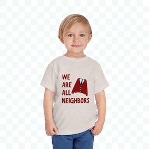 May include: A light beige toddler t-shirt with the text "WE ARE ALL NEIGHBORS" in dark red. A graphic of a red cardigan and a blue tie is printed on the shirt. The child is wearing blue pants.