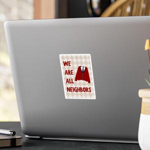 May include: A sticker with the text "WE ARE ALL NEIGHBORS" in red and white. The sticker features a red cardigan with a white shirt and blue tie design. The background is a diamond pattern in beige and white.