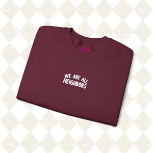 May include: A maroon t-shirt with the white text "WE ARE ALL NEIGHBORS" across the chest. The t-shirt is neatly folded, highlighting the crew neck. The background features a diamond pattern.