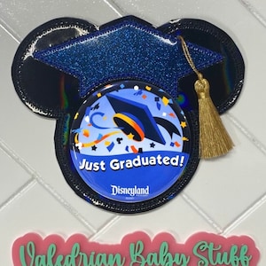 May include: A blue and black glitter graduation cap shaped like Mickey Mouse ears with a gold tassel. The cap has a button with a blue background and a graduation cap with confetti. The button says "Just Graduated!" and "Disneyland Resort". Below the cap is a pink sign that says "Valedrian Baby Stuff & more".