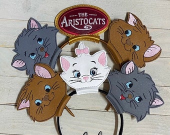 May include: A headband featuring five embroidered cat character heads and a central oval patch with the text THE ARISTOCATS. The cats include a white kitten with a pink bow, two grey kittens, and two brown kittens, all with blue eyes.
