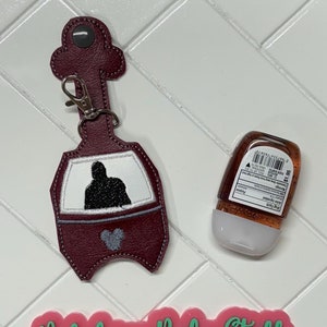 May include: A burgundy fabric hand sanitizer holder with a Mickey Mouse silhouette and a silhouette of a person in a car. The holder has a silver clip and a gray loop. A clear plastic bottle of hand sanitizer is next to the holder. A pink and green sign reads "Valedrian Baby Stuff & more".