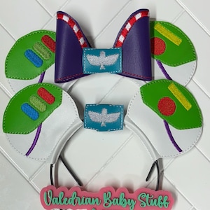 May include: A pair of white headband ears with green and yellow embroidered details, a purple bow with white and red stripes, and a teal blue patch with a white bird design. The headband has a pink sign that says "Valedrian Baby Stuff & more".