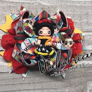 May include: A colorful hair bow with a red, yellow, and blue striped ribbon. The bow features a small doll with black hair, a floral headband, and a black and yellow dress. The doll is holding a yellow scarf. The bow also features a small image of Frida Kahlo. The text "Frida" is visible on the bow.