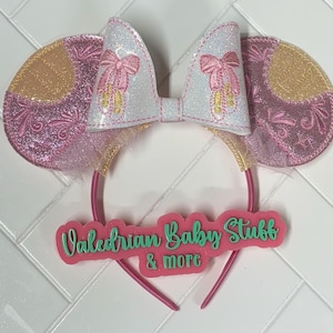 Könnte beinhalten: Rosa und goldene Glitzer-Mausohren mit einer weißen Schleife und einem rosa Haarreif. Der Haarreif hat ein rosa Schild mit der Aufschrift "Valedrian Baby Stuff & more".