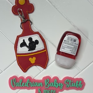 K&ouml;nnte beinhalten: Roter Leder-Schl&uuml;sselanh&auml;nger in Form eines Mickey-Mouse-Fahrzeugs mit einem gelben Mickey-Mouse-Kopf auf der Vorderseite. Der Schl&uuml;sselanh&auml;nger hat einen silbernen Clip und daneben befindet sich eine kleine Flasche Handdesinfektionsmittel. Der Text "Valedrian Baby Stuff & more" befindet sich auf einem rosa Schild unter dem Schl&uuml;sselanh&auml;nger.