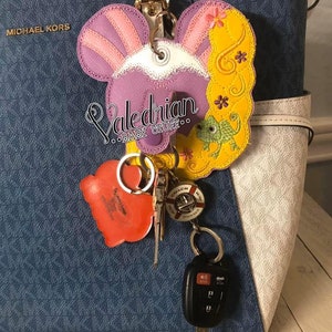 Puede incluir: Un llavero bordado en morado, rosa y amarillo con un dise&ntilde;o de dibujos animados, sujeto a un bolso azul. El llavero presenta un dise&ntilde;o de personaje con el texto "Valedrian BABY STUFFoo". Tambi&eacute;n se adjuntan otras llaves y una etiqueta roja en forma de coraz&oacute;n.