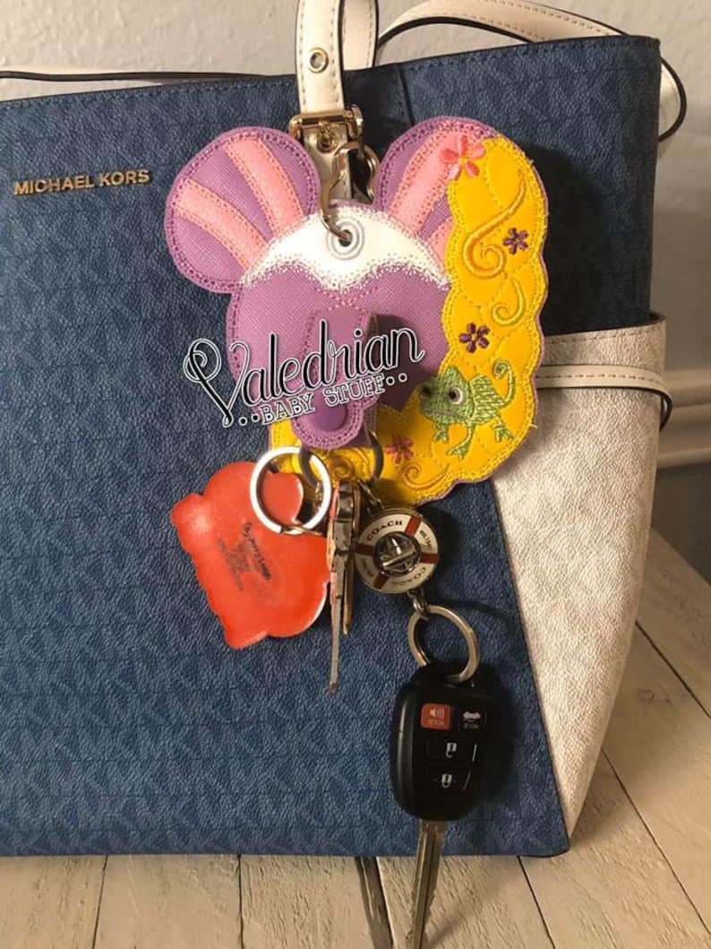 Puede incluir: Un llavero bordado en morado, rosa y amarillo con un dise&ntilde;o de dibujos animados, sujeto a un bolso azul. El llavero presenta un dise&ntilde;o de personaje con el texto "Valedrian BABY STUFFoo". Tambi&eacute;n se adjuntan otras llaves y una etiqueta roja en forma de coraz&oacute;n.