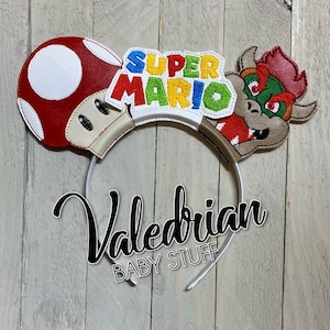 Op de afbeelding: Een witte haarband met Super Mario-thema. Het ontwerp bevat een rood-witte paddenstoel, de tekst "Super Mario" in verschillende kleuren en een Bowser-personage. De tekst "Valedrian BABY STUFF" is ook zichtbaar.