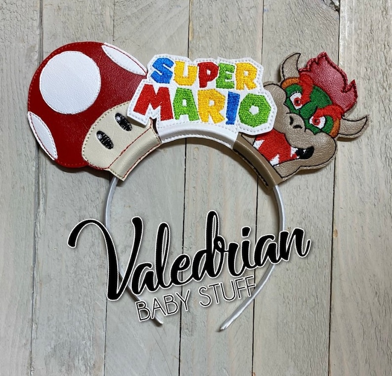 Op de afbeelding: Een witte haarband met Super Mario-thema. Het ontwerp bevat een rood-witte paddenstoel, de tekst "Super Mario" in verschillende kleuren en een Bowser-personage. De tekst "Valedrian BABY STUFF" is ook zichtbaar.