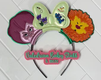 Puede incluir: Una diadema con un lazo verde claro bordado con flores, una abeja y una mecedora. La oreja izquierda es una flor morada oscura con cara, y la derecha es una flor naranja. Un letrero rosa muestra el texto Valedrian Baby Stuff & more.