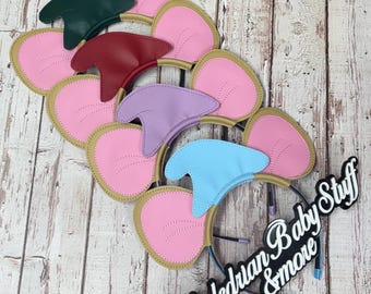 May include: Four headbands featuring pink elephant ears with gold trim and distinctively colored top accents in dark green, red, lavender, and light blue. A black sign with white text reads Valerian Baby Stuff & more.