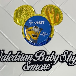 May include: A decorative pin featuring a Minion character from Universal Studios with the text "MY 1ST VISIT." The pin is shaped like Mickey Mouse ears with gold glitter accents. The text "Valedrian Baby Stuff & more" is below the pin.