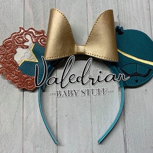 May include: A pair of teal and gold mouse ears with a bow, featuring a brown and gold glitter design on one ear and a teal and gold design with an arrow on the other ear. The text "Valedrian...BABY STUFF..." is printed on the headband.