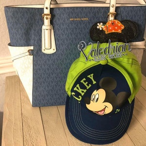 Puede incluir: Un bolso tote azul y blanco con herrajes dorados y el texto "MICHAEL KORS". Una gorra de b&eacute;isbol verde y azul marino con un gr&aacute;fico de Mickey Mouse y la palabra "MICKEY". Una decoraci&oacute;n de oreja de Mickey Mouse negra brillante con una flor.