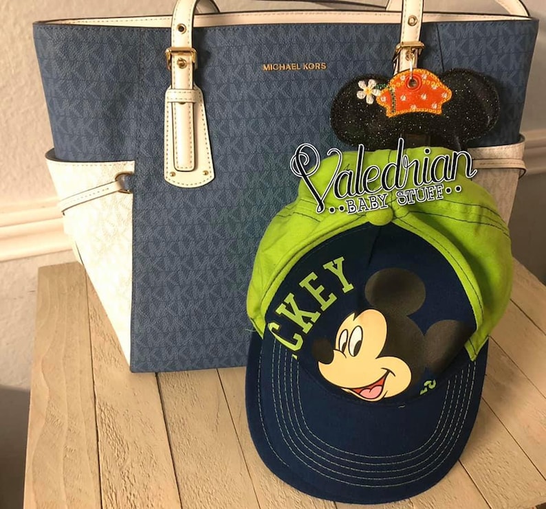 Puede incluir: Un bolso tote azul y blanco con herrajes dorados y el texto "MICHAEL KORS". Una gorra de b&eacute;isbol verde y azul marino con un gr&aacute;fico de Mickey Mouse y la palabra "MICKEY". Una decoraci&oacute;n de oreja de Mickey Mouse negra brillante con una flor.