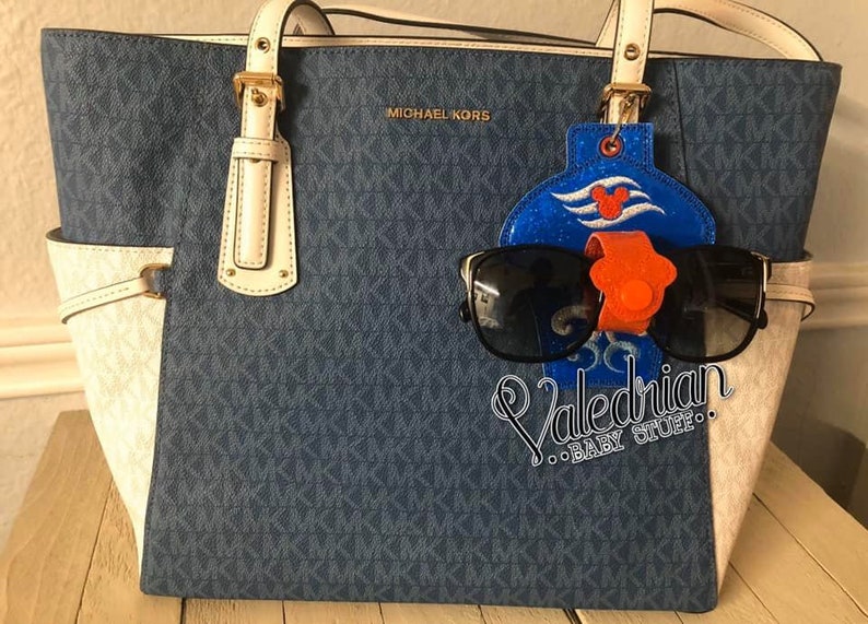 Puede incluir: Un bolso tote azul y blanco con un soporte para gafas de sol y gafas de sol. El bolso tiene un patr&oacute;n repetitivo y las palabras "MICHAEL KORS" son visibles. El soporte para gafas de sol es azul con un dise&ntilde;o de Mickey Mouse y el texto "Valedrian BABY STUFF".