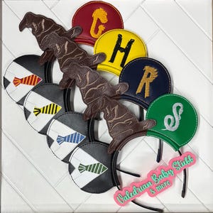 May include: A collection of themed headbands featuring various house emblems and Sorting Hats. The headbands have black bands with round, two-tone ear designs. The Sorting Hats are brown, and the house emblems are in red, yellow, navy, and green.