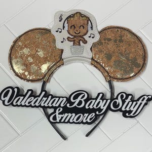 May include: A pair of mouse ears with a brown and gold sequined design. The center features a white applique of a cartoon character wearing headphones. The text "Valedrian Baby Stuff & more" is displayed in a black and white font.