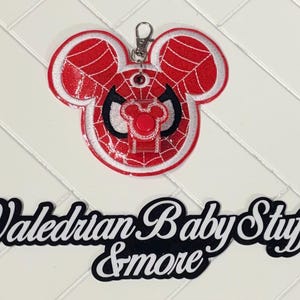 Puede incluir: Un llavero con la cabeza de Mickey Mouse roja y blanca con el tema de Spider-Man. El llavero tiene un cierre plateado y el texto "Valedrian Baby Stuff & more" en blanco y negro.
