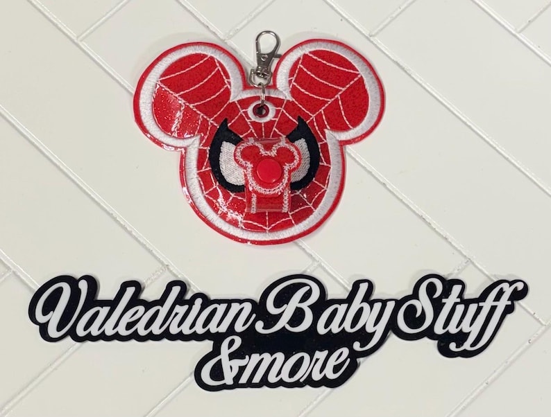 Puede incluir: Un llavero con la cabeza de Mickey Mouse roja y blanca con el tema de Spider-Man. El llavero tiene un cierre plateado y el texto "Valedrian Baby Stuff & more" en blanco y negro.