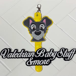 May include: A yellow lanyard with a cartoon dog face design. The dog has gray fur, pink ears, and a black nose. The lanyard has a metal clasp and the text "Valedrian Baby Stuff & more" in black and white.