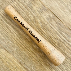May include: A wooden cocktail muddler with the text "Cocktail Queen!" in black. The muddler is cylindrical with a rounded end and a small string around the middle. The background is a light brown wood grain.
