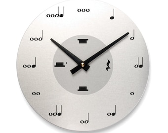 May include: A round, silver-colored wall clock with black musical note hour markers. The clock face has a light gray center circle. Black hour and minute hands are positioned at 2:10. A gold-colored dot is at the center.