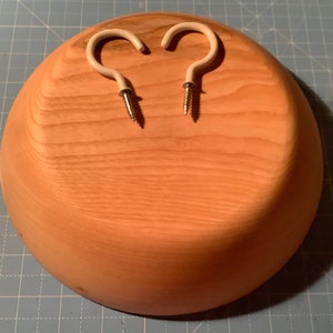 May include: A wooden bowl with two white hooks attached to the bottom. The bowl is light brown and has a smooth finish.