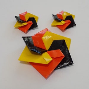 Puede incluir: Un conjunto de tres pendientes de origami en negro, amarillo y naranja. Los pendientes tienen forma de flores con cinco pétalos.
