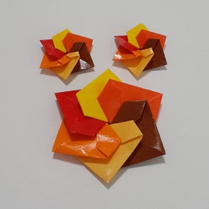 Puede incluir: Un conjunto de joyas de origami, que incluye un colgante y pendientes a juego. Las piezas en forma de estrella presentan una vibrante paleta de colores rojo, naranja, amarillo y marrón. Las joyas se exhiben sobre un fondo blanco.
