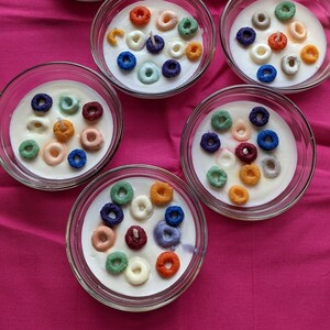 May include: Seven clear glass bowls filled with white wax, each decorated with colorful cereal pieces and a small candle. The bowls are arranged on a bright pink surface, creating a playful and unique decorative item.