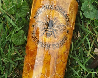 May include: A yellow and gold tumbler with a black bee and the text "Be the change you wish to see" in a circle around the bee. The tumbler has a honeycomb pattern and a dripping honey effect.