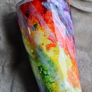 May include: A stainless steel tumbler with a rainbow-colored design. The tumbler has a clear lid and is decorated with a swirling pattern of red, orange, yellow, green, blue, and purple. The design is accented with gold glitter.