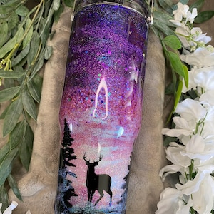 May include: A cylindrical tumbler with a black lid and handle, featuring a glittery gradient design that transitions from deep purple at the top to pink and light blue at the base, with a black silhouette of a deer and pine trees.