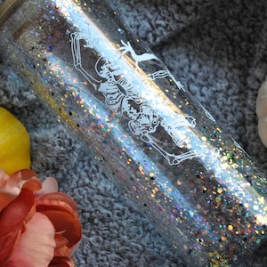 May include: A clear glass tumbler with a glittery iridescent finish and a white skeleton design. The tumbler is sitting on a gray fuzzy surface.