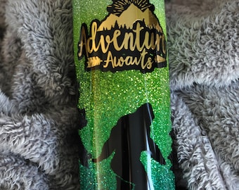 Può includere: Un bicchiere verde e nero con glitter con il testo "Adventure Awaits" e una silhouette di montagna. Il bicchiere ha una silhouette nera di una persona che scia in discesa.