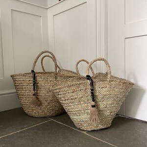 May include: Two large woven straw baskets with black leather handles and black tassel accents. The baskets are sitting on a gray tiled floor.
